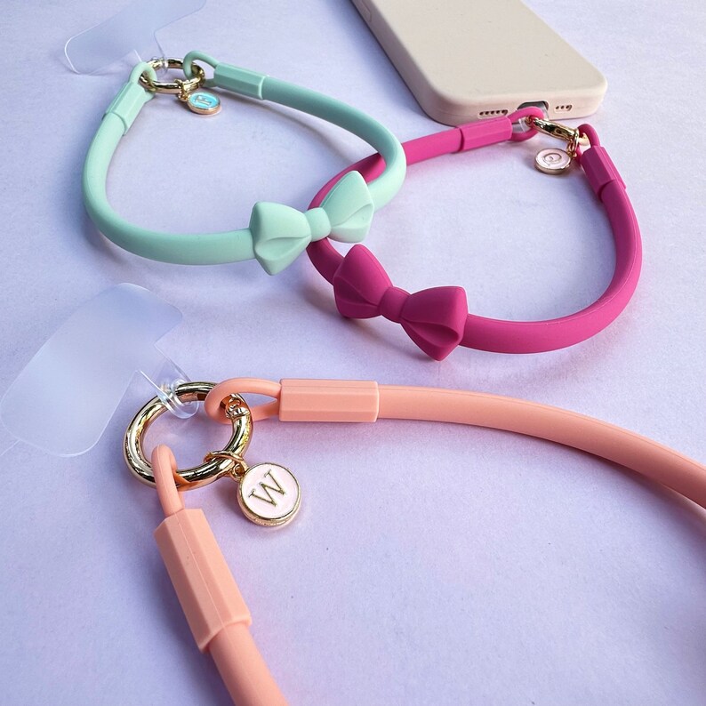 May include: Three silicone phone straps with bow accents. The straps are in mint green, pink, and peach. Each strap has a gold charm with a letter on it. The charms are "W", "A", and a small circle.