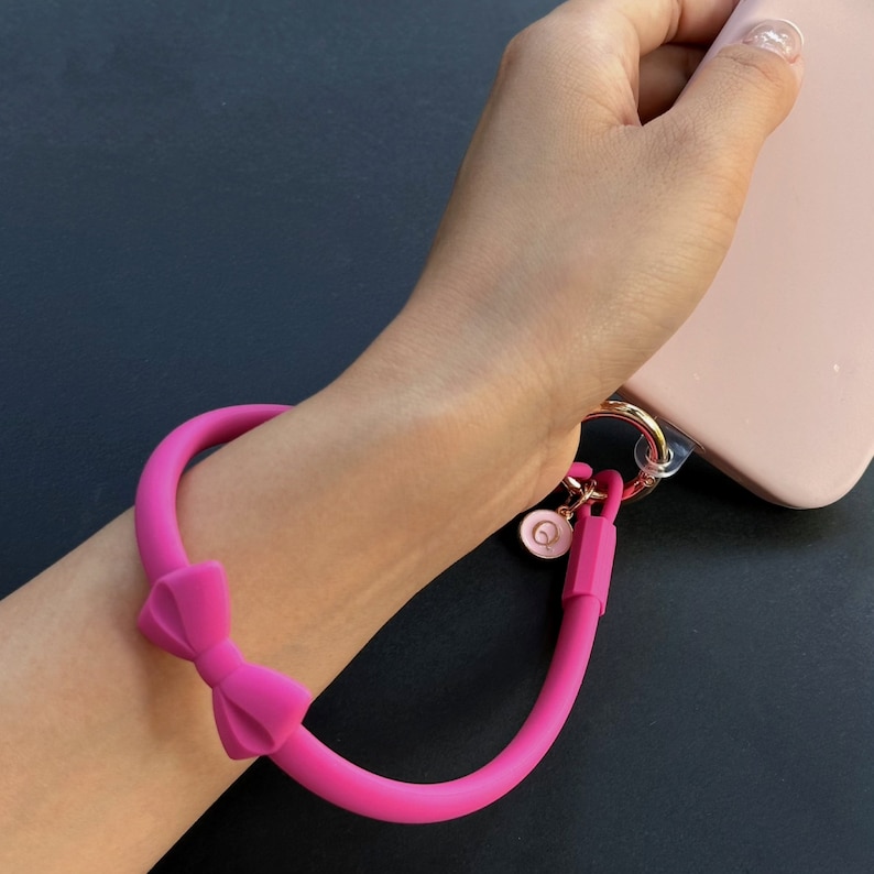May include: A pink silicone phone strap with a bow detail and a gold charm. The strap is attached to a phone case.
