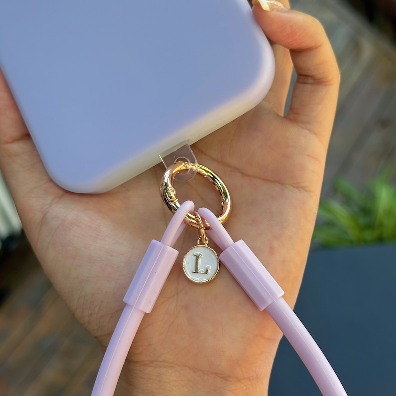 May include: A lavender phone case with a clear strap and a gold charm with a white letter L.