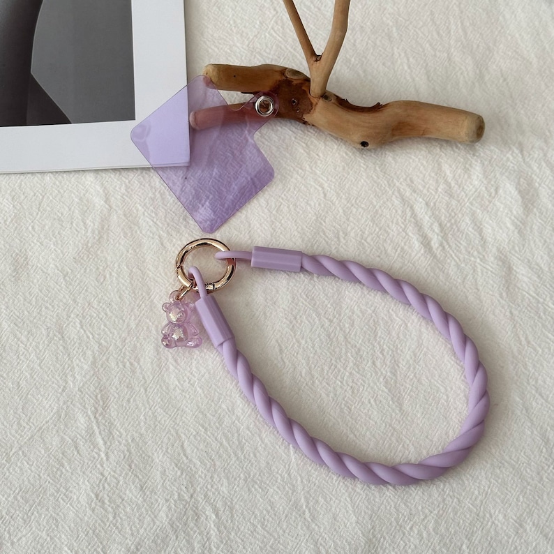 May include: A lavender-colored phone strap with a twisted rope design and a small, clear, lavender-colored bear charm. The strap has a gold-colored metal ring.