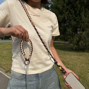 May include: A brown and cream braided phone strap and matching wristlet. The strap is attached to a clear phone case. The strap is worn across the body. The wristlet is held in the hand.