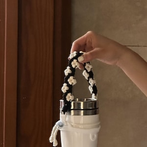 May include: A white and black crocheted handle for a water bottle. The handle is made of a thick cord and has a flower pattern.