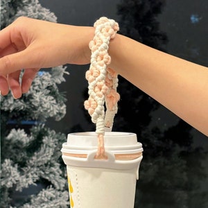 May include: A pink and white crocheted cup holder with a flower design. The holder is attached to a white to-go cup.