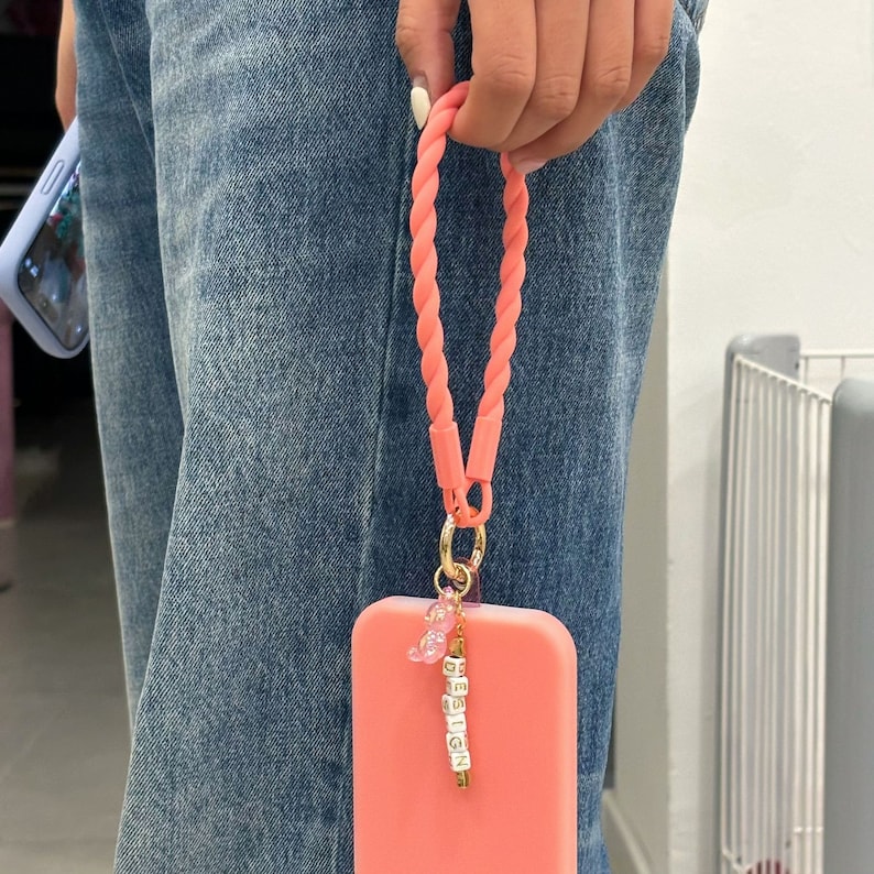 May include: A coral pink phone case with a matching coral pink braided phone strap. The strap has a gold charm with the word "DESIGN" spelled out in white beads.
