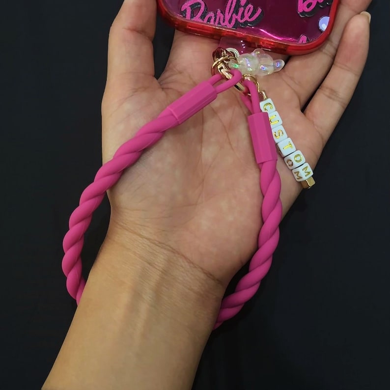 May include: A pink phone case with a pink braided phone strap and a charm that says "CUSTOM".