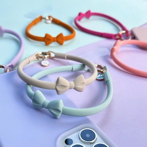 May include: A set of four silicone phone straps with bow accents. The straps are in various colours, including light blue, beige, orange, and pink.
