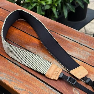 May include: A camera strap featuring a woven green and white diagonal pattern on the exterior and a solid black padded interior. The strap includes light tan leather end tabs with visible stitching and black adjustable nylon webbing connectors.