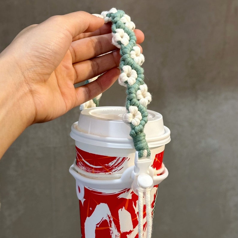 May include: A white and green crocheted cup holder with white flowers. The holder is attached to a red and white cup with a black design.