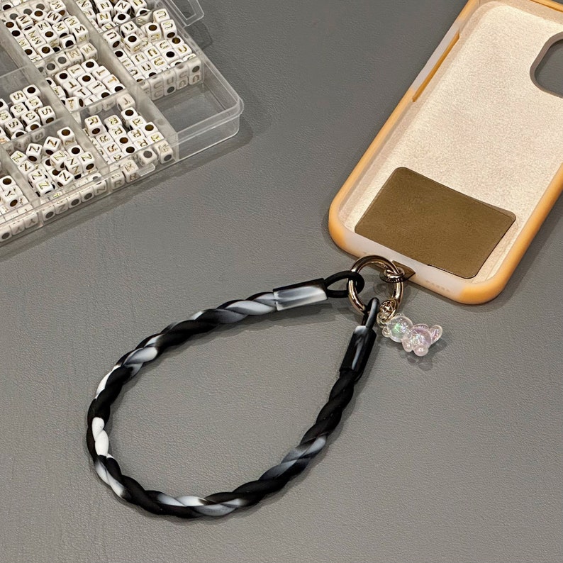 May include: A black and white braided phone strap with a gold ring and a clear iridescent bear charm attached. The strap is connected to a light brown phone case.