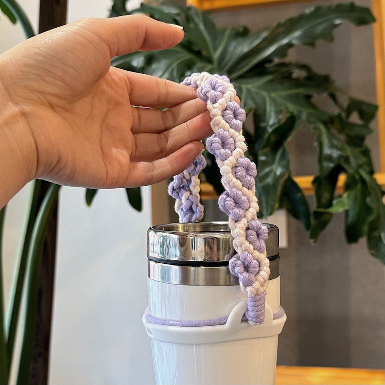 May include: A white and purple crocheted handle for a reusable water bottle. The handle is made of a soft, textured yarn and has a flower design.