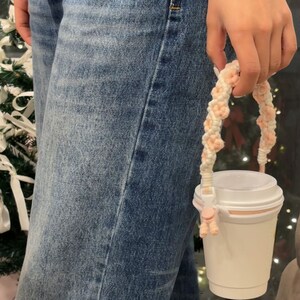 May include: A white to-go cup with a pink and white braided handle. The handle is attached to the cup with a pink plastic clip.
