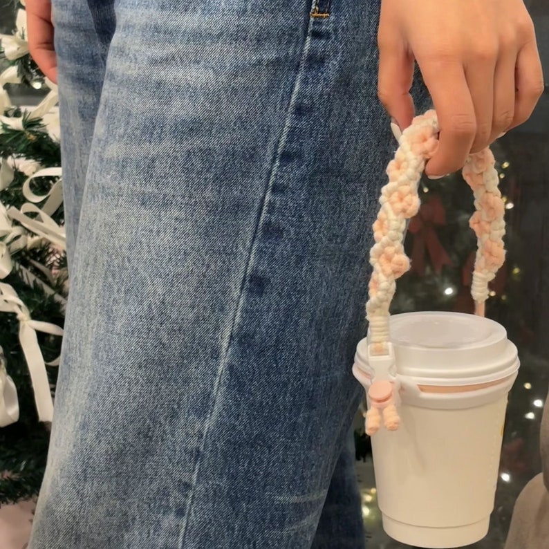 May include: A white to-go cup with a pink and white braided handle. The handle is attached to the cup with a pink plastic clip.