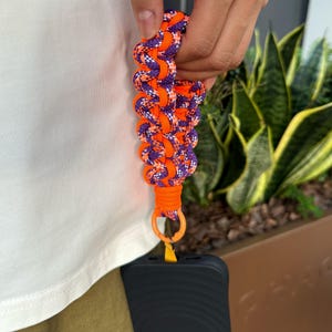 May include: A purple and orange braided phone strap with a black phone case attached. The strap has a metal ring and a yellow cord.