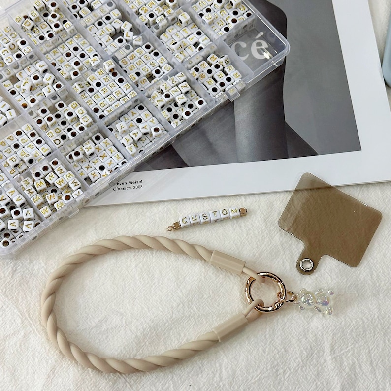 May include: A beige rope phone strap with a gold ring and a small white bear charm. The strap is next to a container of white letter beads with gold lettering. The beads spell out "CUSTOM".