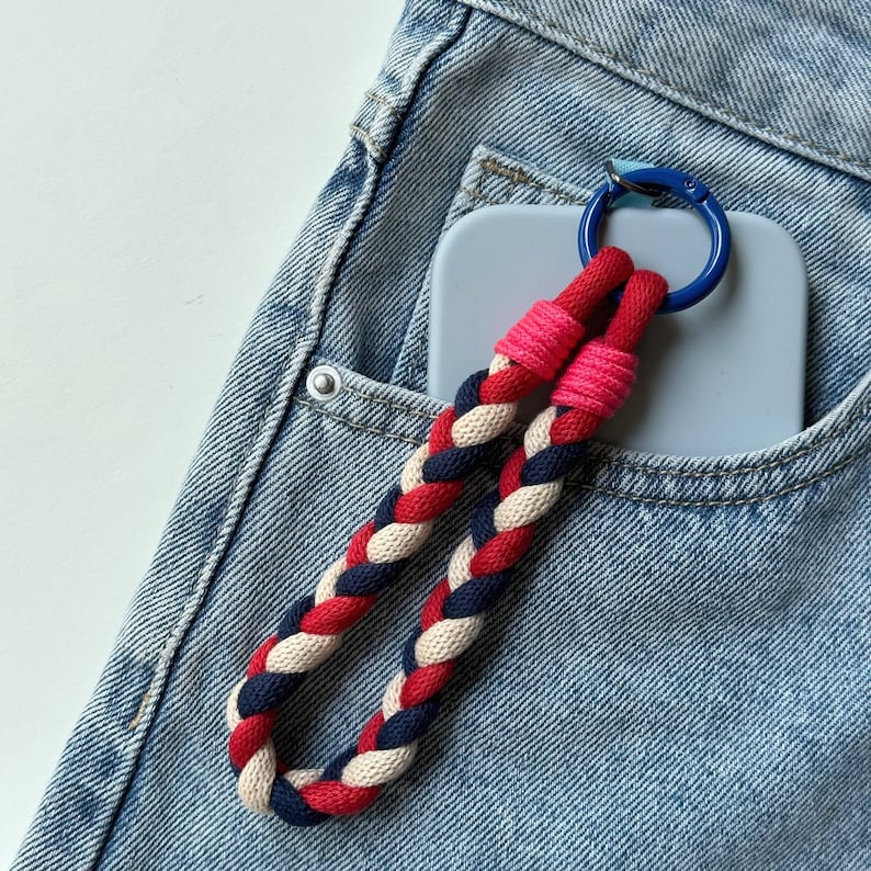K&ouml;nnte beinhalten: Ein blaues und wei&szlig;es Handy-Case mit einem rot-wei&szlig;-blauen geflochtenen Seil-Handyband, das daran befestigt ist. Das Handy-Case und das Band befinden sich in einer blauen Jeans-Tasche.