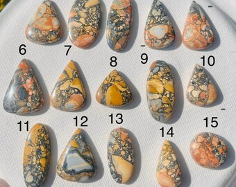 May include: Fifteen polished gemstone cabochons in various shapes, including oval, teardrop, and triangular. The stones feature a mix of colors, including orange, yellow, gray, and black, with intricate patterns. The gemstones are displayed on a white surface.