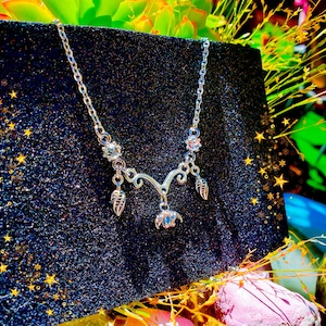 May include: A silver-toned necklace with a delicate design. The necklace features a central floral pendant, two leaf charms, and sparkling accents. The chain is displayed against a dark, glittery background, with star-shaped embellishments.
