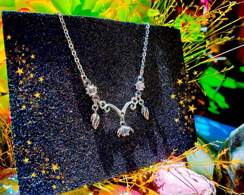 May include: A silver-toned necklace with a delicate design. The necklace features a central floral pendant, two leaf charms, and sparkling accents. The chain is displayed against a dark, glittery background, with star-shaped embellishments.