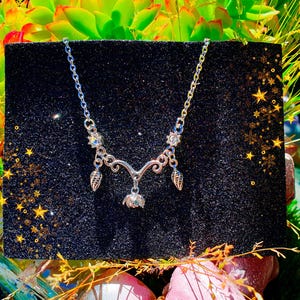 May include: A silver-toned necklace with a delicate floral and leaf design. The necklace features a central pendant and two leaf-shaped charms. The necklace is displayed on a black, glittery surface with a backdrop of green plants and gold stars.