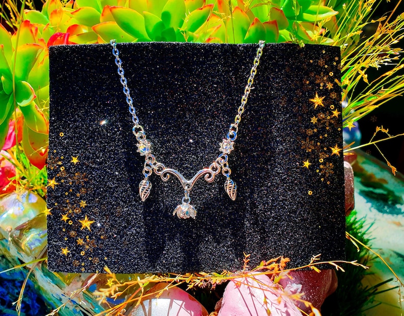 May include: A silver-toned necklace with a delicate floral and leaf design. The necklace features a central pendant and two leaf-shaped charms. The necklace is displayed on a black, glittery surface with a backdrop of green plants and gold stars.