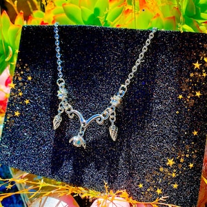 May include: A silver-toned necklace with a delicate floral and leaf design. The necklace is displayed on a black, glittery surface, with a backdrop of green and yellow foliage. The necklace features a chain and several decorative elements.