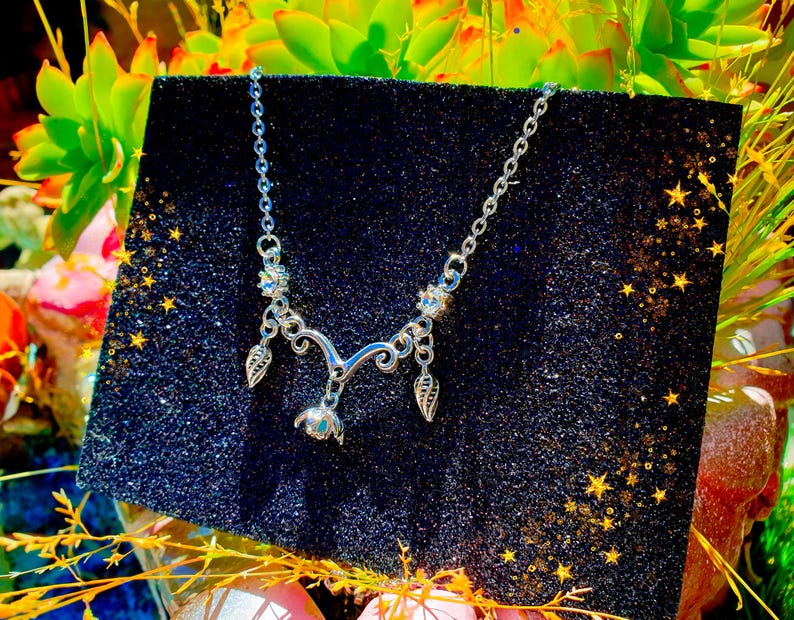 May include: A silver-toned necklace with a delicate floral and leaf design. The necklace is displayed on a black, glittery surface, with a backdrop of green and yellow foliage. The necklace features a chain and several decorative elements.