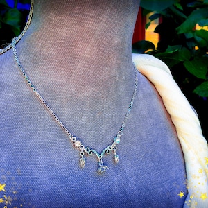 May include: Silver-toned necklace with a delicate design, featuring a central pendant and smaller charms. The necklace is displayed on a mannequin with a blue fabric background and a cream-colored scarf. The image has a soft, artistic aesthetic.