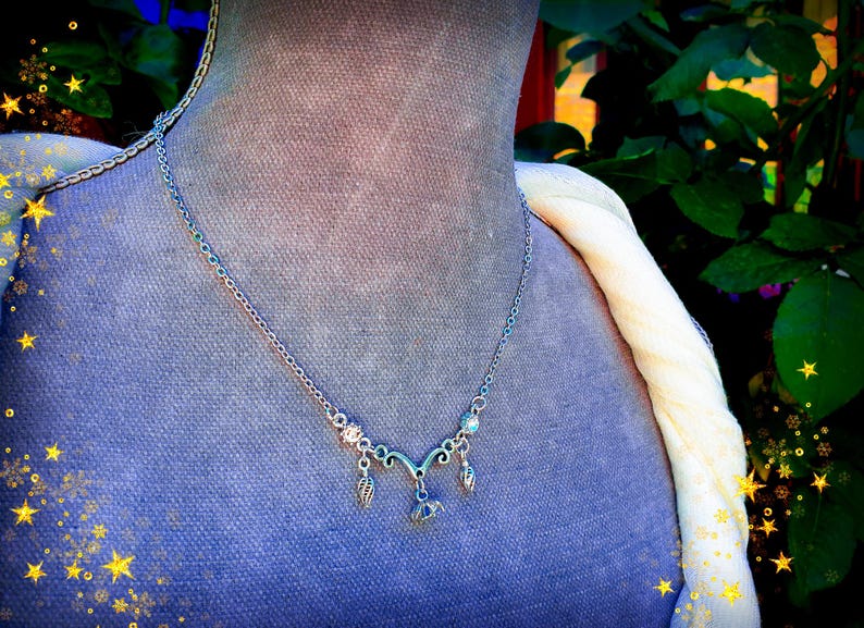 May include: Silver-toned necklace with a delicate design, featuring a central pendant and smaller charms. The necklace is displayed on a mannequin with a blue fabric background and a cream-colored scarf. The image has a soft, artistic aesthetic.