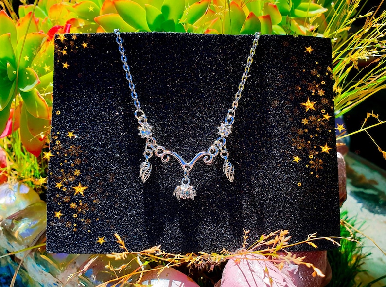 May include: A silver-toned necklace with a floral pendant and leaf-shaped charms. The necklace is displayed on a black, glittery surface, surrounded by greenery and gold star accents. The necklace has a delicate, feminine design.