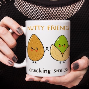 May include: White ceramic mug with a cartoon almond and pistachio holding hands. The mug reads "Nutty Friends cracking smiles".