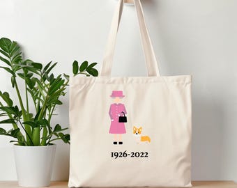 Könnte beinhalten: Eine weiße Canvas-Tragetasche mit einer rosa und schwarzen Illustration von Queen Elizabeth II und einem Corgi. Der Text "1926-2022" ist unter der Illustration gedruckt.