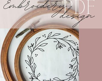Może przedstawiać: Drewniana tamborki do haftu z kwiatowym wzorem. Wzór to czarny rysunek linii wieńca z liści i jagód na białej tkaninie. Słowa "Embroidery PDF design" są napisane kursywą.