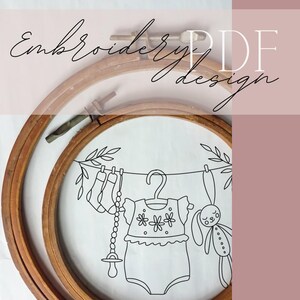 Op de afbeelding: Een borduurring met een babythema-ontwerp. Het ontwerp omvat een romper, sokken, een fopspeen en een knuffelkonijn dat aan een waslijn hangt. De woorden "Embroidery PDF design" zijn cursief geschreven.