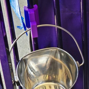 Op de afbeelding: Een glanzende roestvrijstalen emmer met een gebogen handvat. De emmer is bevestigd aan een paars frame met een paarse plastic clip. Een blauwe plastic zak is zichtbaar op de achtergrond. De emmer is ontworpen om vloeistoffen of andere materialen in te bewaren.
