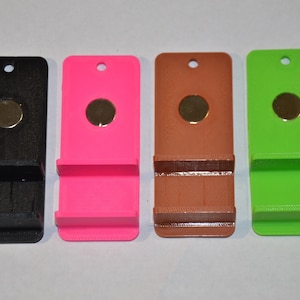 May include: Four phone stands in different colors: black, pink, brown, and green. Each stand has a small round magnet on the front and a slot for a phone.
