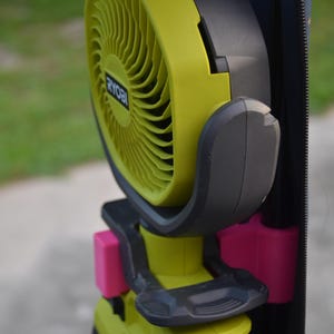 May include: A yellow and black Ryobi fan with a pink clip attached to a black bag. The fan is powered by a battery pack.