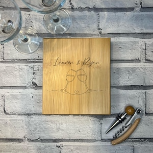May include: A personalised bamboo chopping board engraved with the names "Lauren & Ryan". The board features a design of two wine glasses clinking together with a heart in the middle. A wooden corkscrew is also pictured.