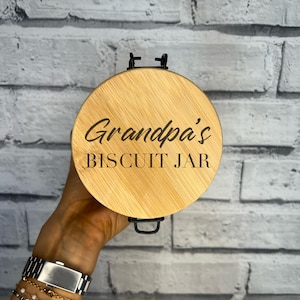 May include: A circular wooden lid, light in colour, engraved with "Grandpa's BISCUIT JAR" in a dark font. It features a black metal handle and is set against a grey brick wall.