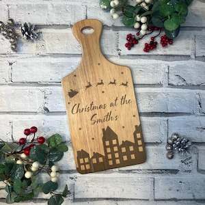 May include: A wooden chopping board with a Christmas theme. It features a silhouette of Santa Claus flying over a city skyline with the text "Christmas at the Smith's".