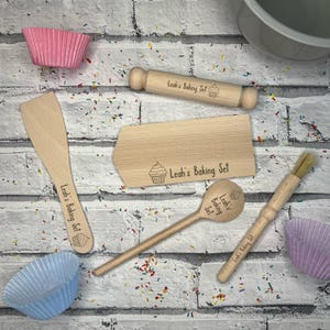 May include: A wooden baking set featuring the text "Leah's Baking Set" and a cupcake design. The set includes a rolling pin, spatula, scraper, spoon, and brush. Pink and blue paper baking cups are also visible.
