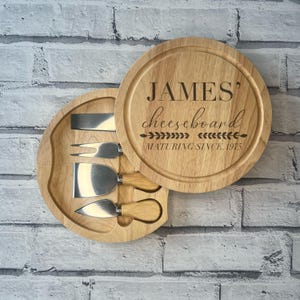 May include: A round wooden cheeseboard with a handle and four cheese knives. The board is engraved with the text "JAMES' cheeseboard MATURING SINCE 1975".
