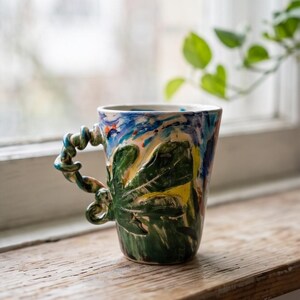 May include: A ceramic mug featuring a hand-painted surface with blue, yellow, and green abstract brushstrokes. The exterior displays a raised, sculpted green leaf design. The handle is uniquely shaped like a twisted, coiled vine in shades of blue and green.