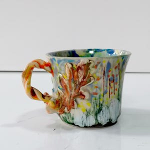 May include: A ceramic mug featuring a multi-colored, abstract drip-paint design in shades of blue, green, yellow, and orange. The mug includes a three-dimensional sculpted oak leaf and acorn detail on the side, with a twisted, multi-colored handle.