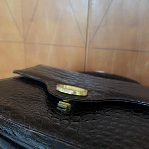 Puede incluir: Bolso de mano negro texturizado con un cierre dorado. El bolso tiene un cierre de solapa y un acabado brillante. El material parece tener un patr&oacute;n de piel de cocodrilo o de caim&aacute;n.