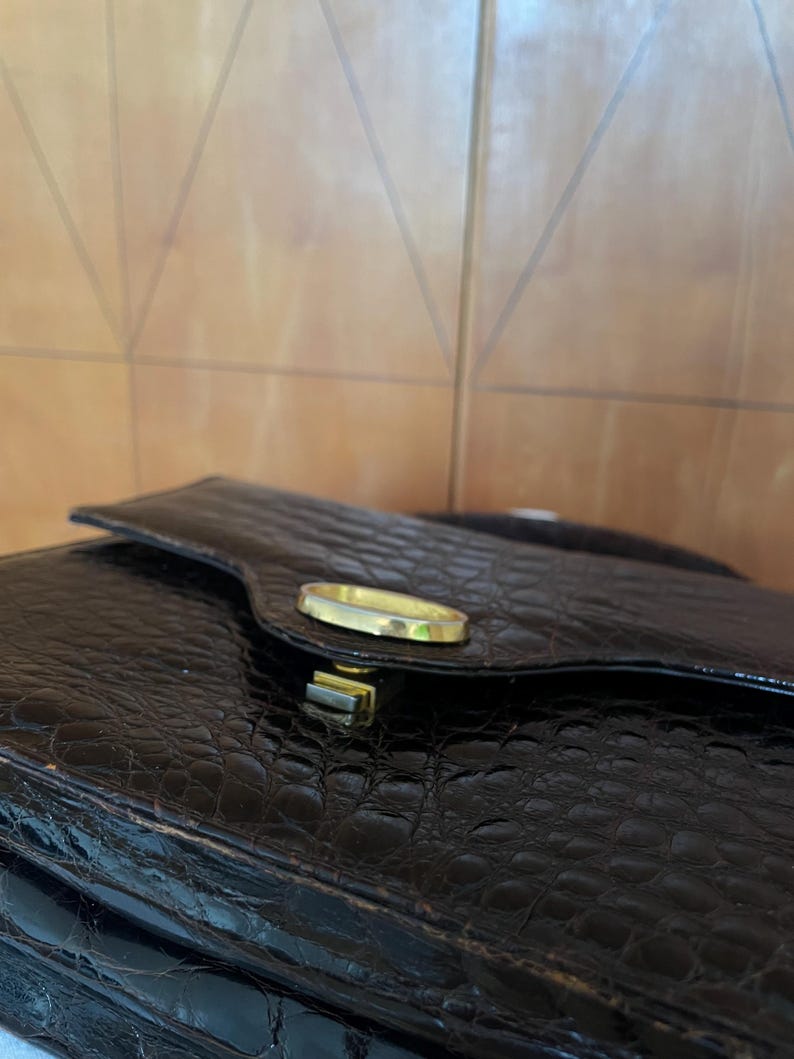 Puede incluir: Bolso de mano negro texturizado con un cierre dorado. El bolso tiene un cierre de solapa y un acabado brillante. El material parece tener un patr&oacute;n de piel de cocodrilo o de caim&aacute;n.