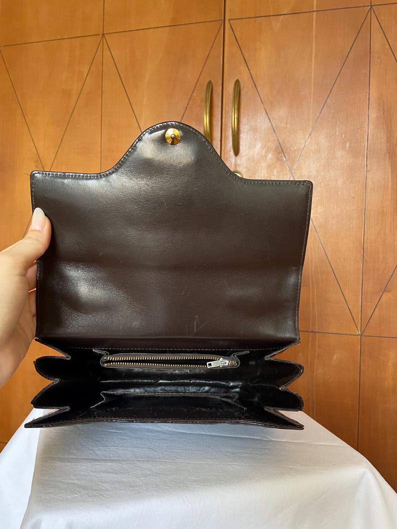 Puede incluir: Bolso de mano de cuero marr&oacute;n oscuro, abierto para mostrar el interior. Cuenta con un cierre dorado y un dise&ntilde;o estructurado con m&uacute;ltiples compartimentos. El interior incluye un bolsillo con cremallera y forro marr&oacute;n oscuro.
