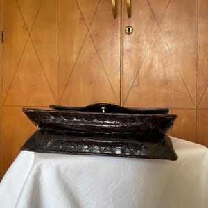 Puede incluir: Bolso de mano marr&oacute;n oscuro de estilo vintage con un patr&oacute;n texturizado similar al de un cocodrilo. El bolso presenta un cierre dorado y un dise&ntilde;o estructurado. El bolso est&aacute; sobre una superficie blanca.