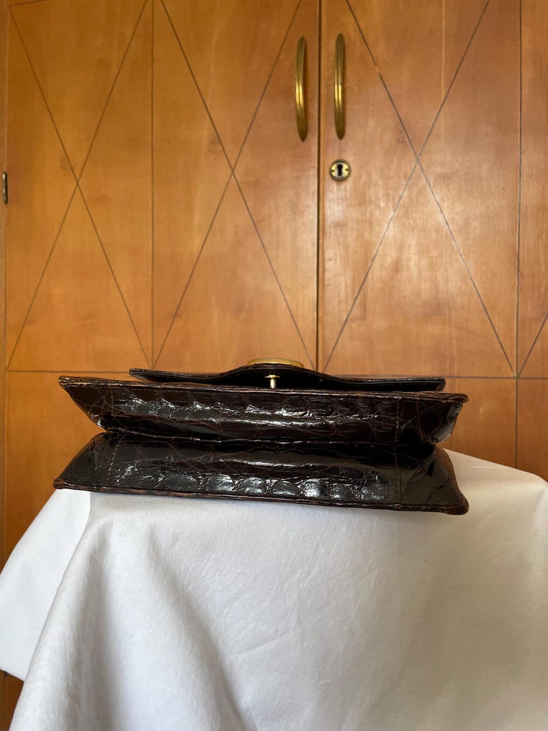 Puede incluir: Bolso de mano marr&oacute;n oscuro de estilo vintage con un patr&oacute;n texturizado similar al de un cocodrilo. El bolso presenta un cierre dorado y un dise&ntilde;o estructurado. El bolso est&aacute; sobre una superficie blanca.