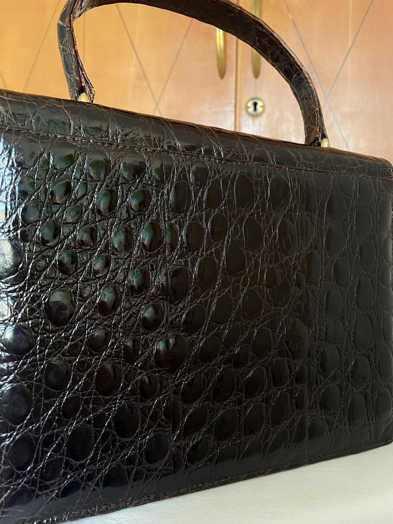 Puede incluir: Un bolso vintage marr&oacute;n oscuro con un patr&oacute;n texturizado similar al de un cocodrilo. El bolso tiene un asa superior y una forma rectangular. La superficie tiene un acabado brillante y los herrajes parecen ser dorados.