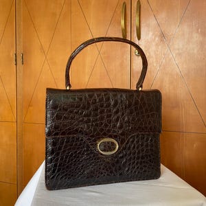 Puede incluir: Un bolso vintage marr&oacute;n con un patr&oacute;n texturizado similar al de un cocodrilo. El bolso tiene un asa curva, un cierre ovalado dorado y detalles dorados. El bolso est&aacute; sobre una superficie blanca.